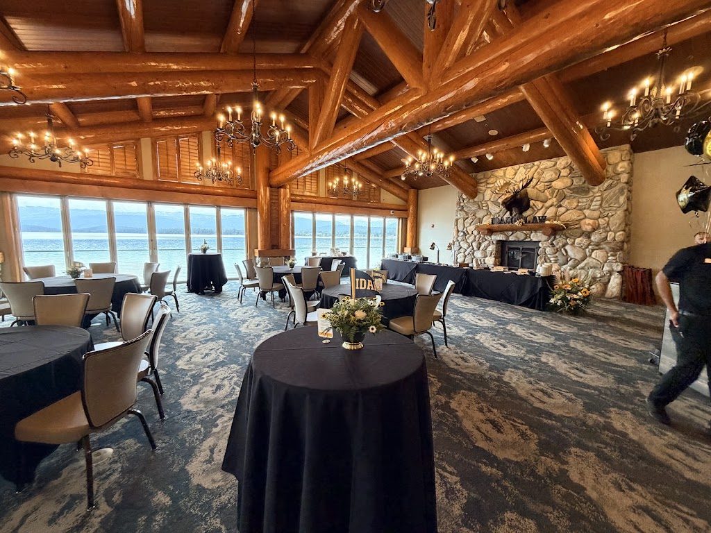 A room with log beams, chandeliers, a stone fireplace and lake windows